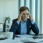 Ein gestresster Büroangestellter sitzt an einem Schreibtisch mit Unterlagen und Laptop und reibt sich erschöpft die Schläfen.
